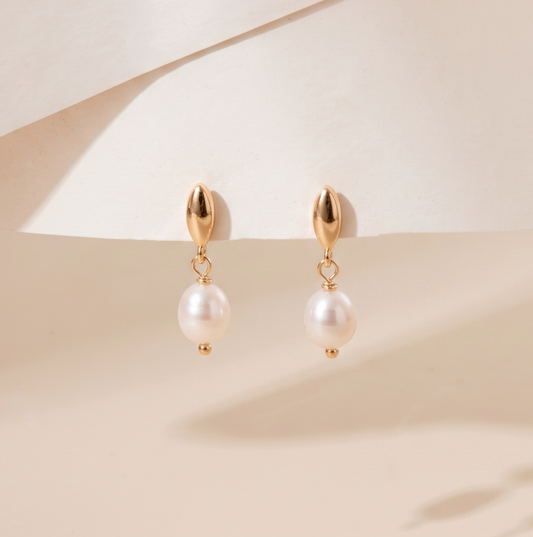 A pair of gold drop earrings with white pearl-like beads, displayed on a soft beige surface with elegant shadows.