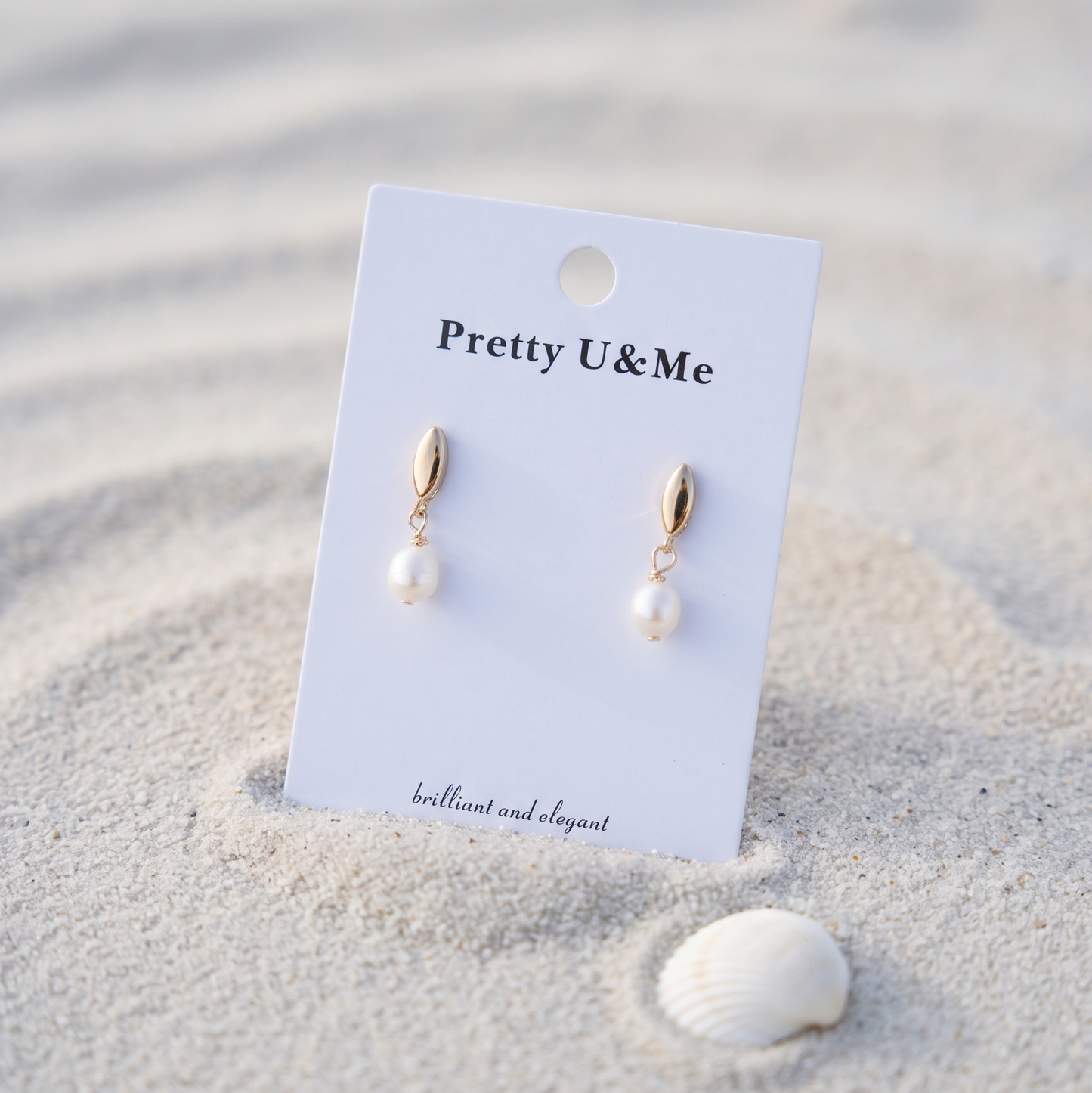 Gold drop earrings with white pearl pendants displayed on a white card labeled 'Pretty U&Me', set in sand next to a small seashell.