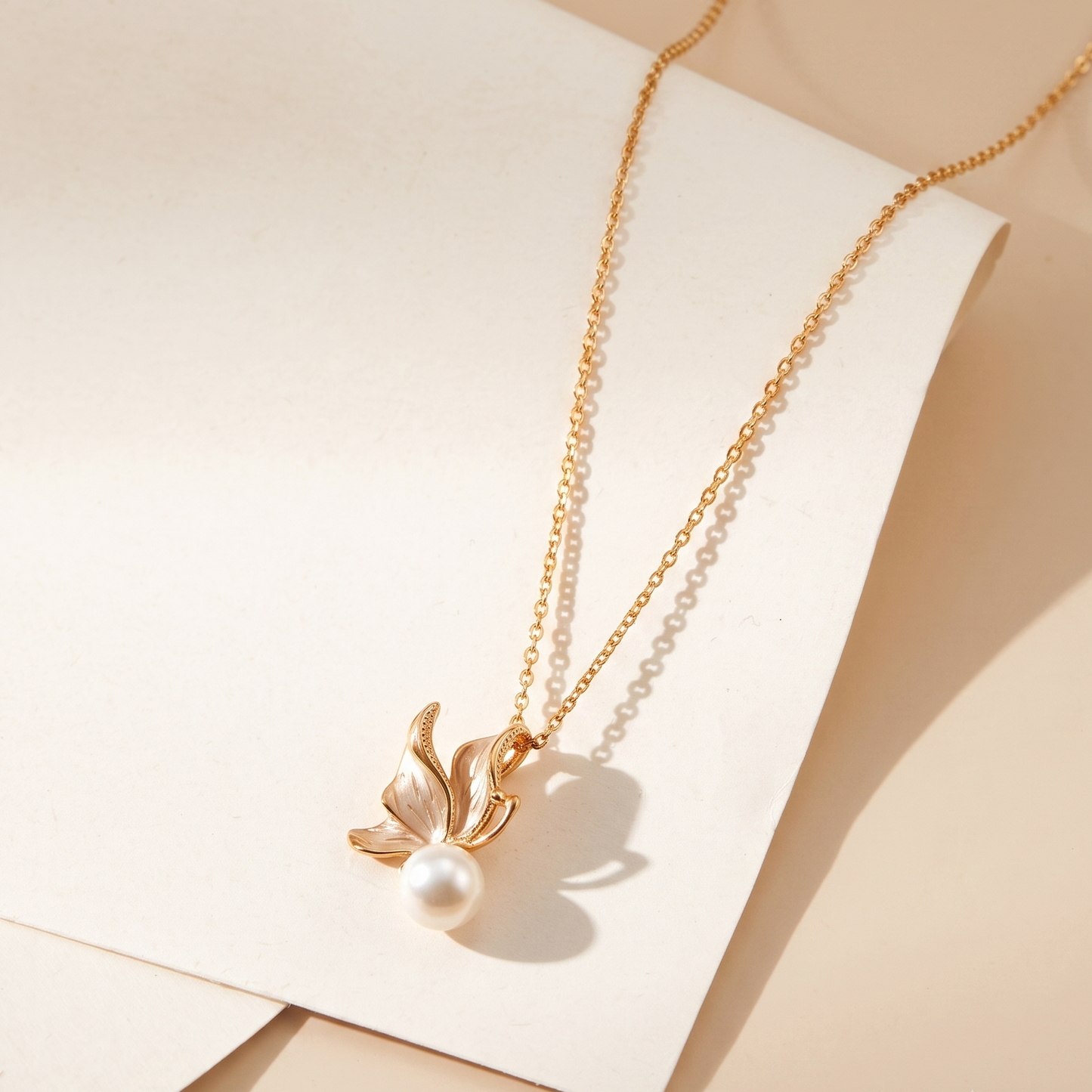A gold chain necklace with a delicate gold leaf pendant and a single white pearl, displayed on a cream-colored card against a warm beige background.