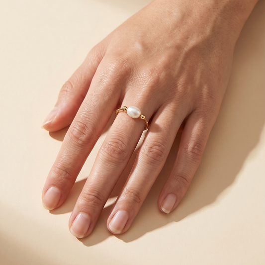 A delicate gold ring with a small white pearl center, worn on a person's ring finger against a warm beige background.