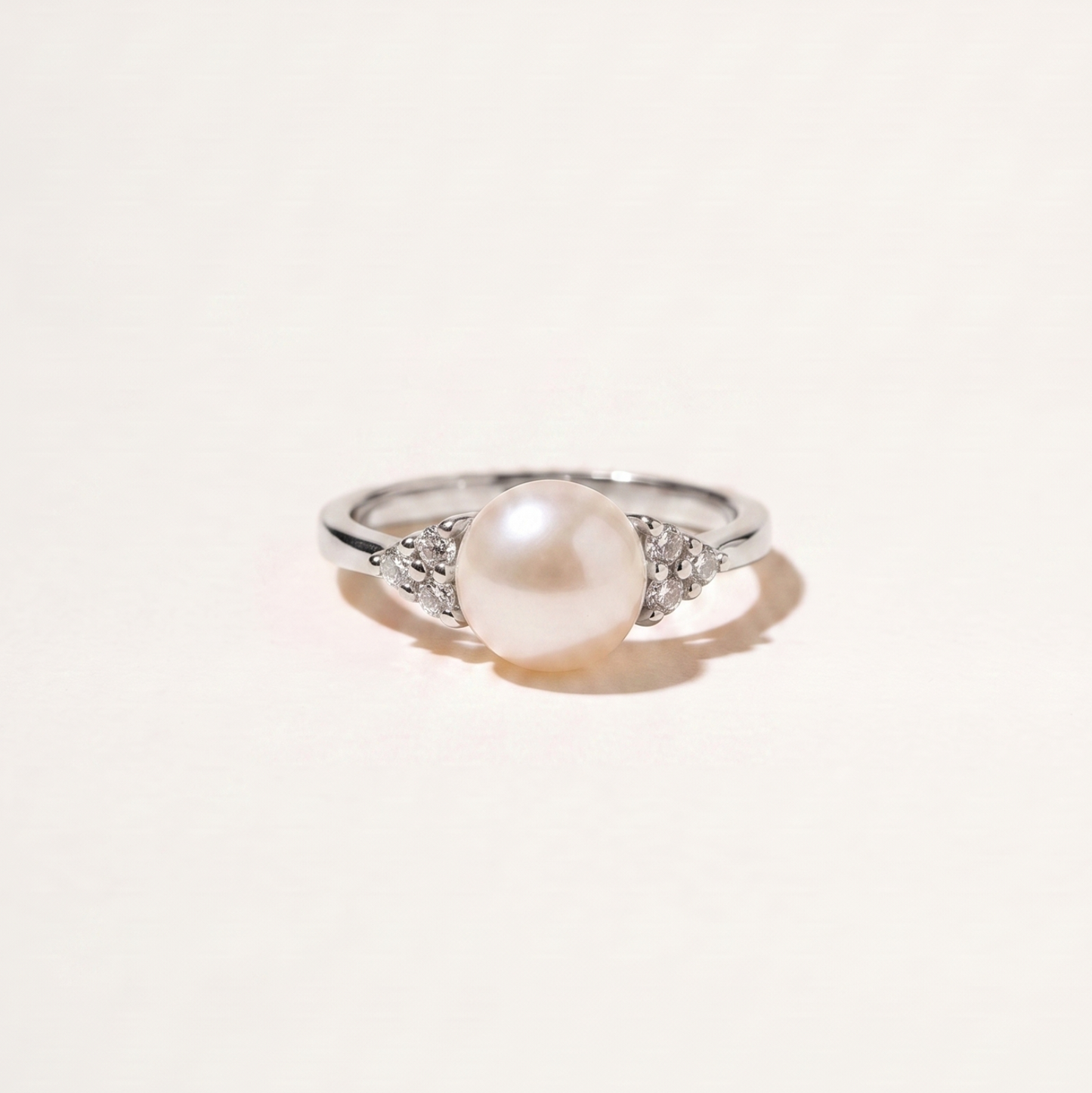 A silver ring featuring a central white pearl and small clear stones on either side of the band, photographed on a plain off-white background.