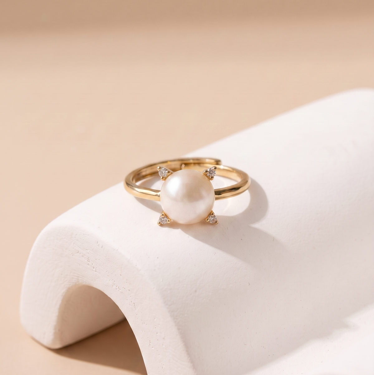 A gold ring with a large white pearl and small clear stones, displayed on a white curved jewelry stand against a warm beige background.