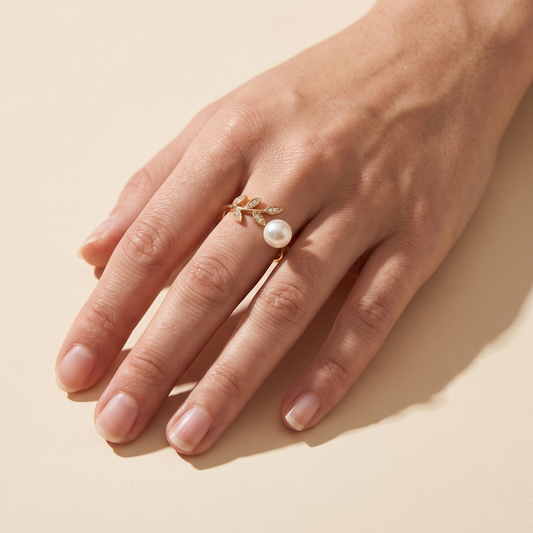 A close-up of a hand wearing a delicate gold ring with a small white pearl, set against a warm beige background.