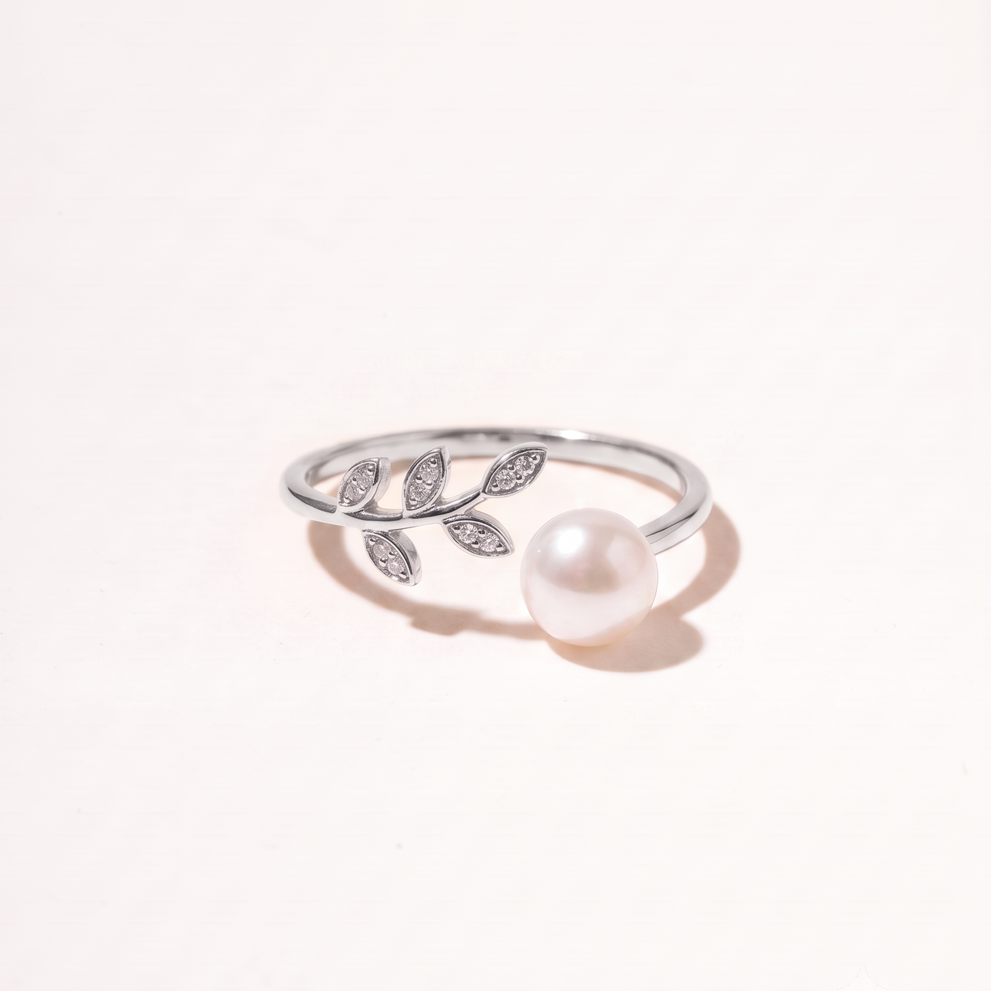 A silver leaf ring with a single white pearl, photographed on a clean, light beige background.