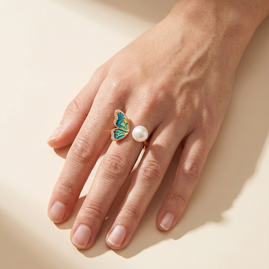 A close-up of a hand wearing a gold butterfly ring with green and blue enamel details and a white pearl, set against a warm beige background with soft shadows.