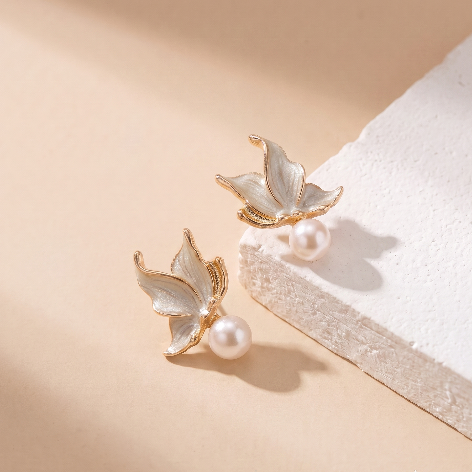 A pair of gold and white enamel butterfly stud earrings with pearl accents, displayed on a textured white block against a warm beige background.