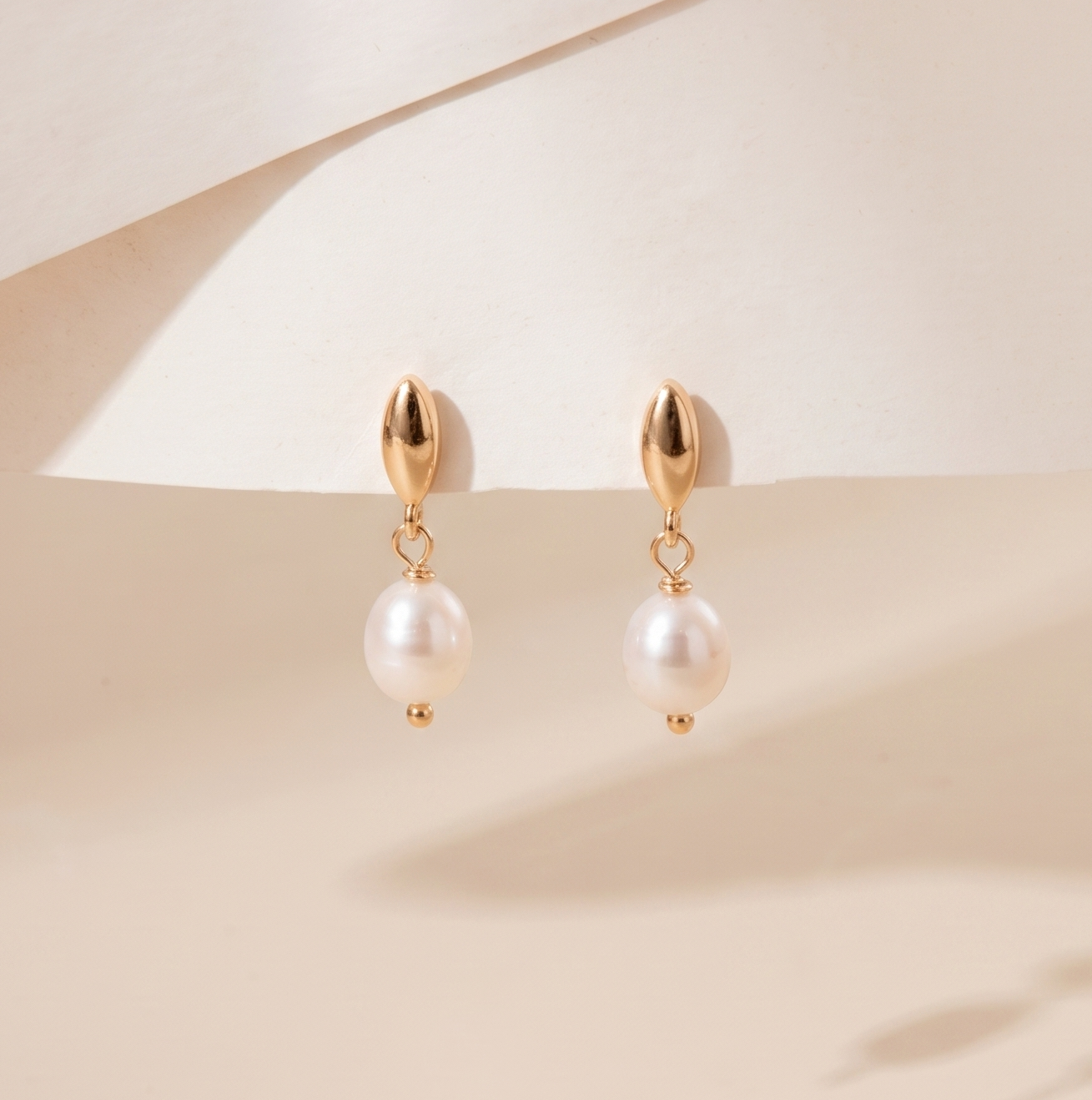 A pair of gold drop earrings with white pearl-like beads, displayed on a soft beige surface with elegant shadows.