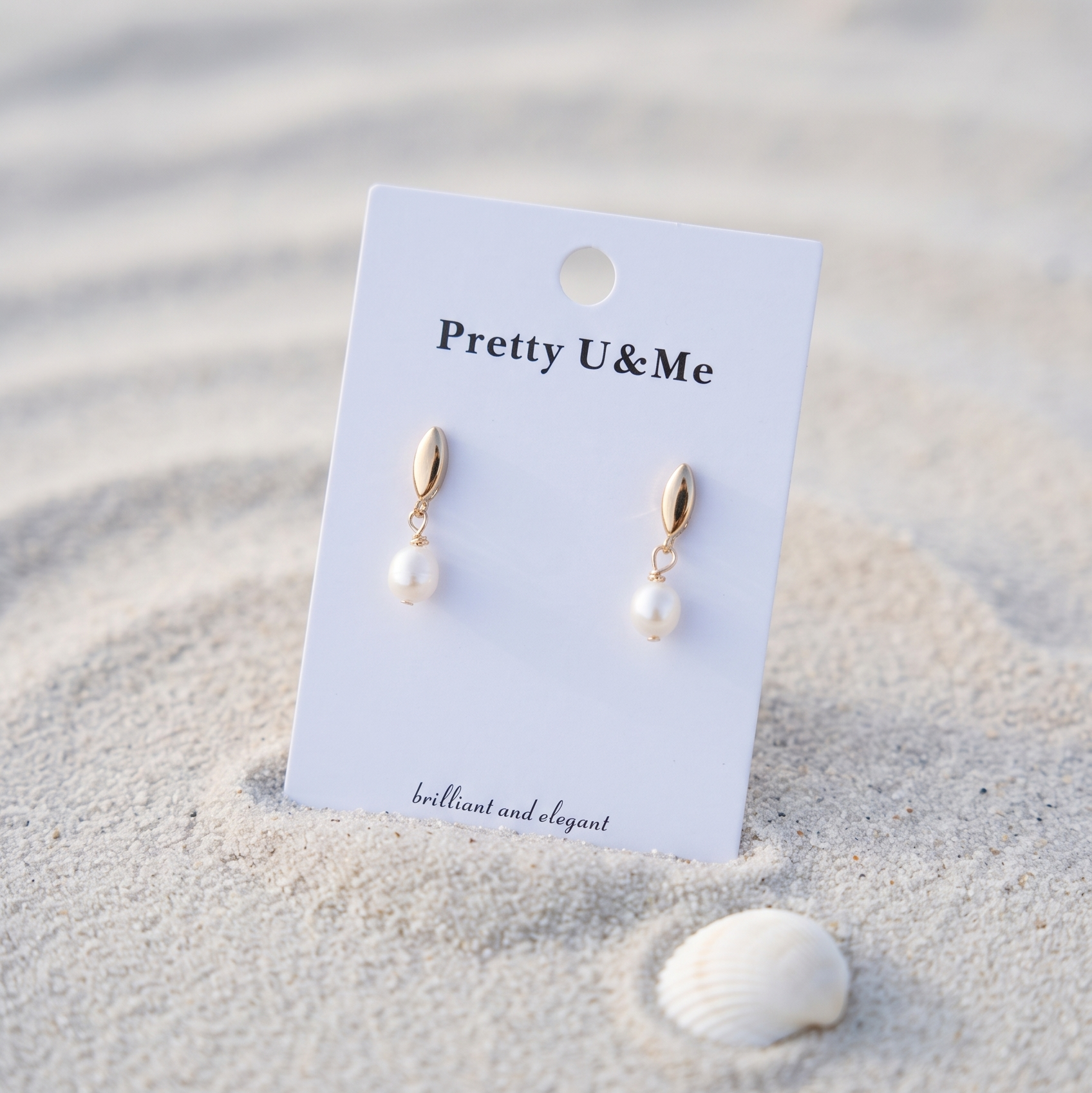 Gold drop earrings with white pearl pendants displayed on a white card labeled 'Pretty U&Me', set in sand next to a small seashell.
