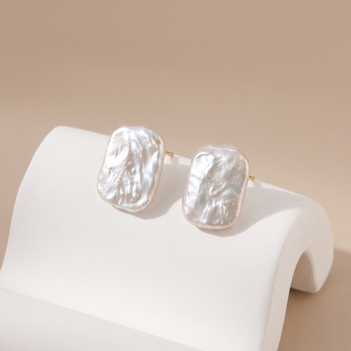 Two textured white pearl stud earrings with gold posts, displayed on a white curved jewelry stand against a warm beige background.