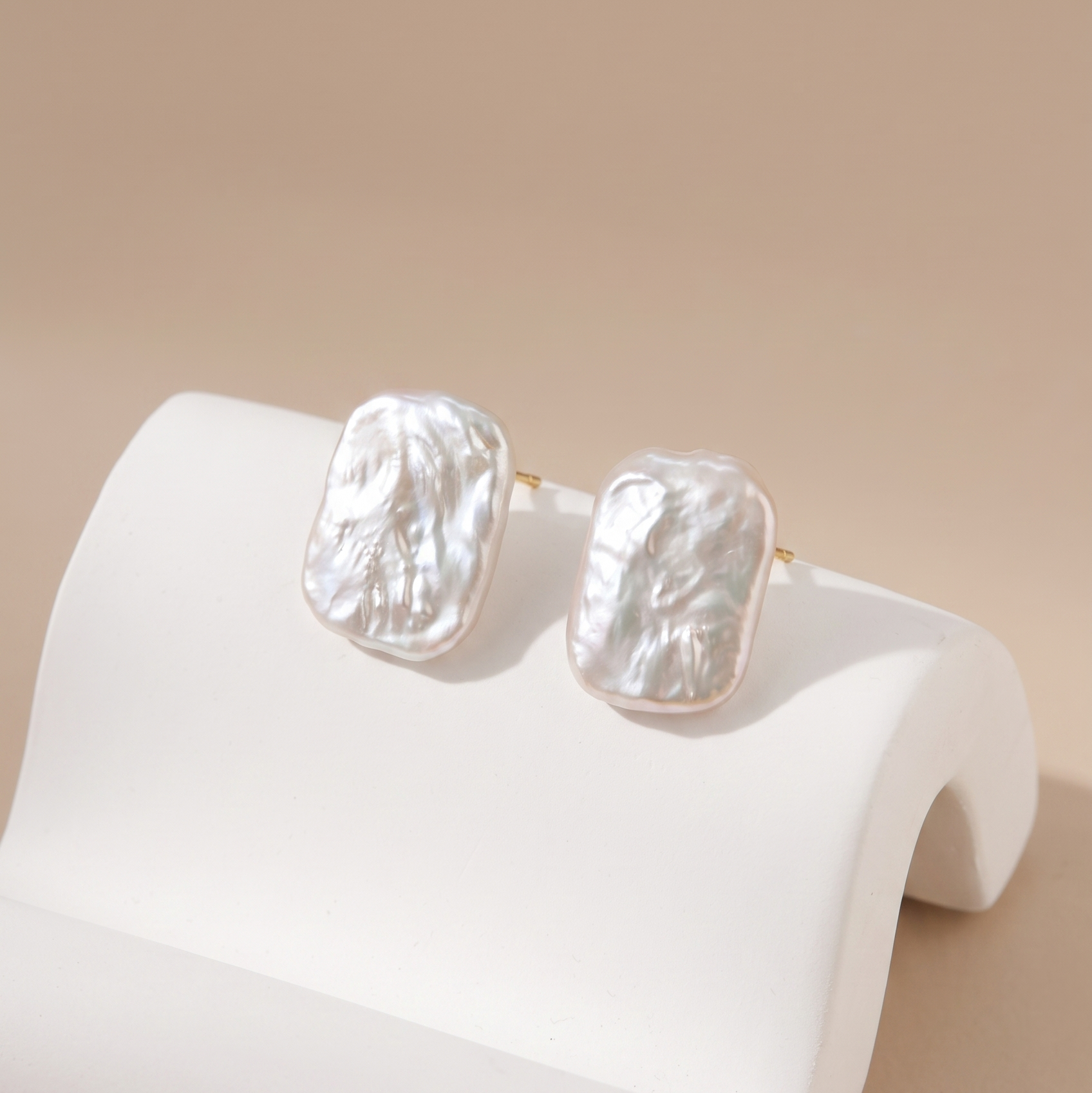 Two textured white pearl stud earrings with gold posts, displayed on a white curved jewelry stand against a warm beige background.