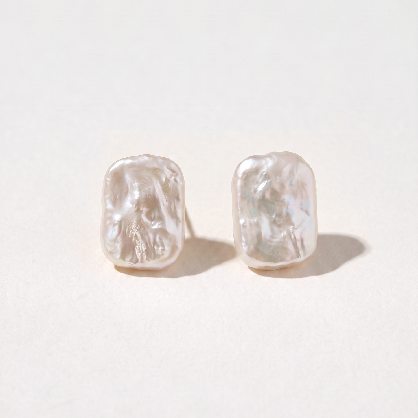 A pair of textured, organic-shaped stud earrings in a warm off-white or pale pinkish-beige tone, displayed on a clean, light cream background.