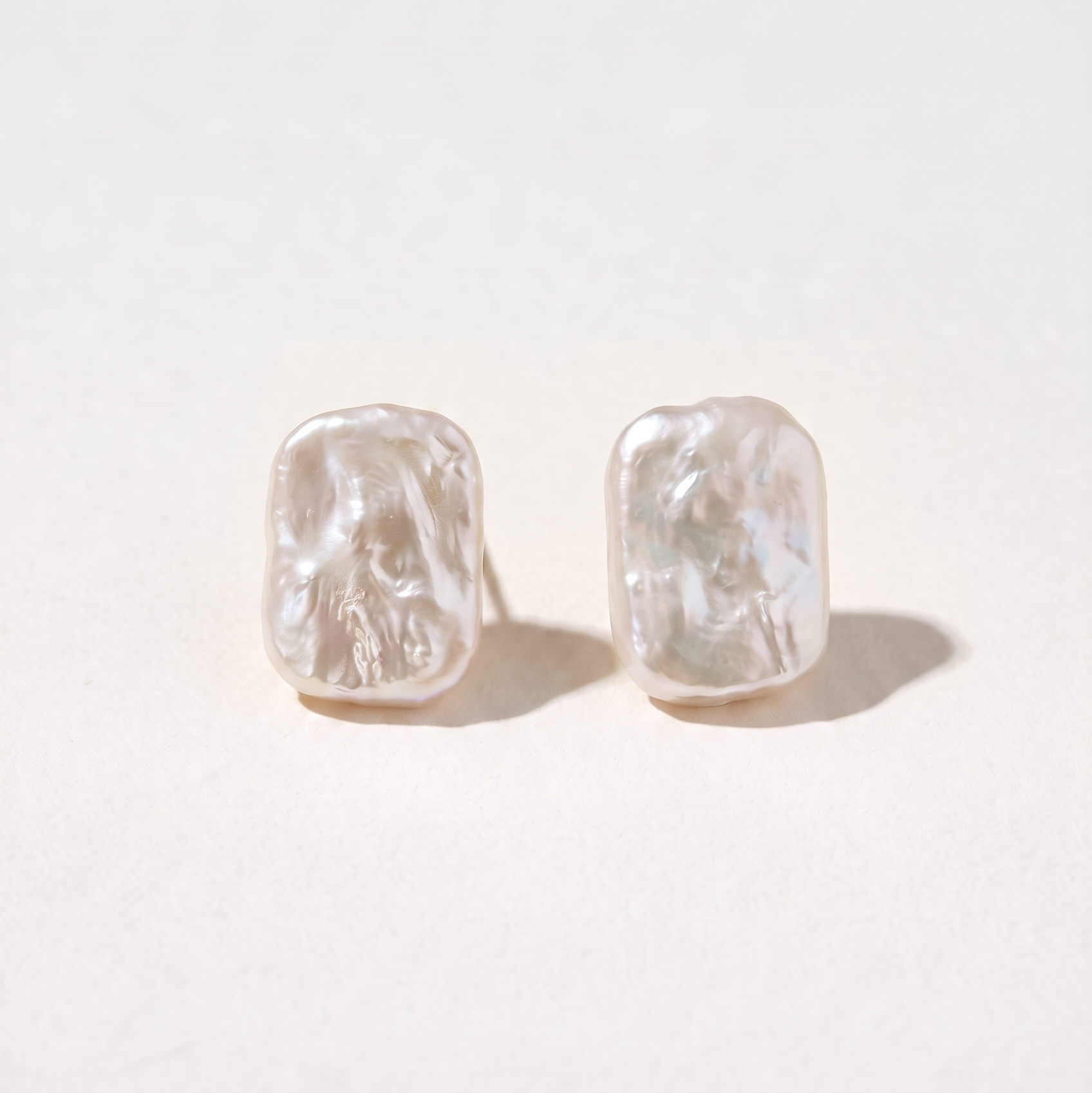 A pair of textured, organic-shaped stud earrings in a warm off-white or pale pinkish-beige tone, displayed on a clean, light cream background.