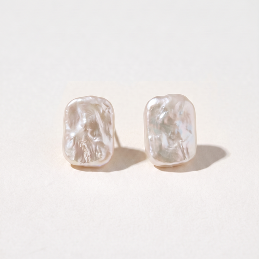 A pair of textured, organic-shaped stud earrings in a warm off-white or pale pinkish-beige tone, displayed on a clean, light cream background.