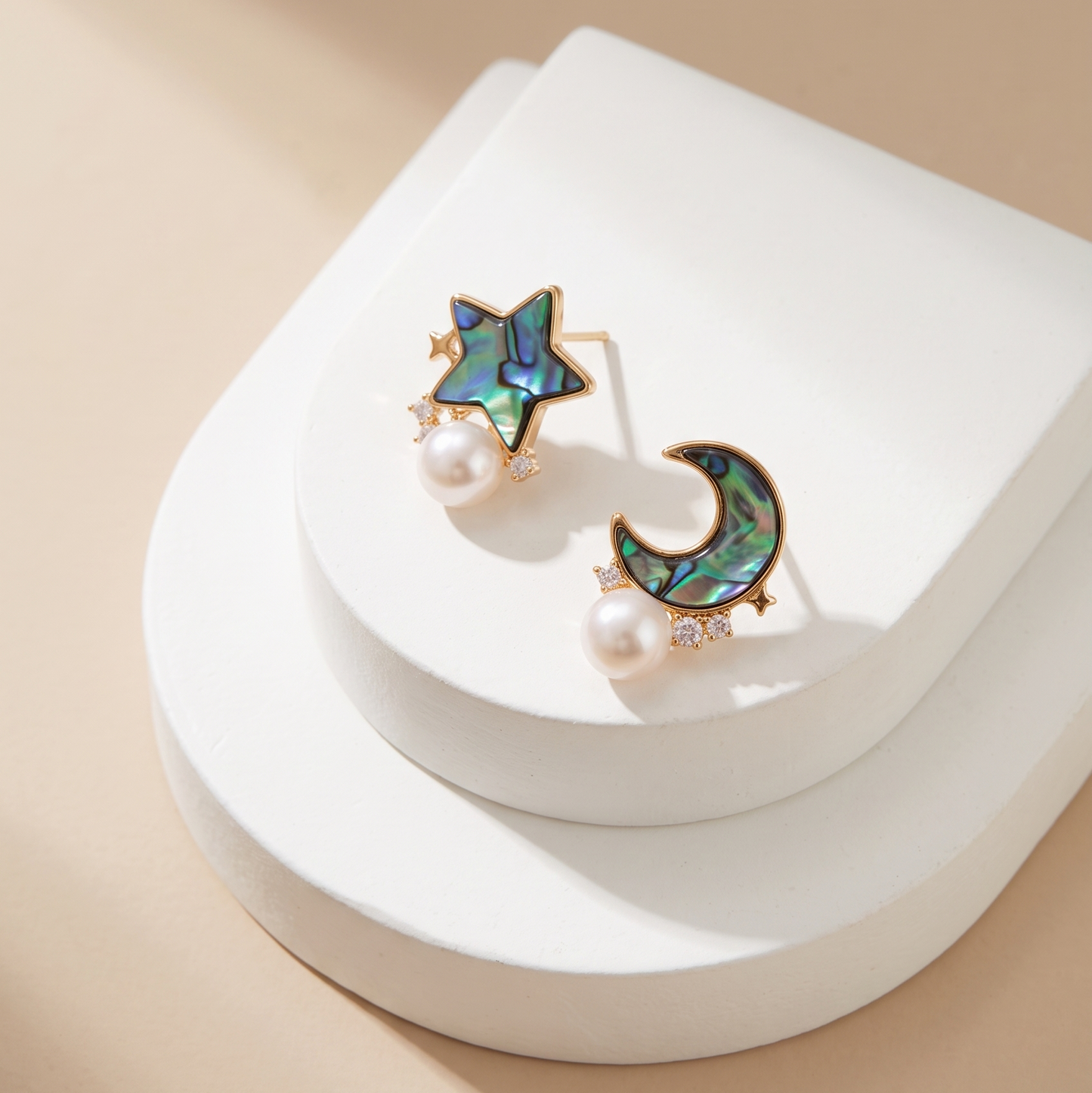 A pair of celestial stud earrings displayed on a white tiered jewelry stand. The earrings feature a gold-tone star and crescent moon design with iridescent abalone shell inlay and a small white pearl hanging below each.