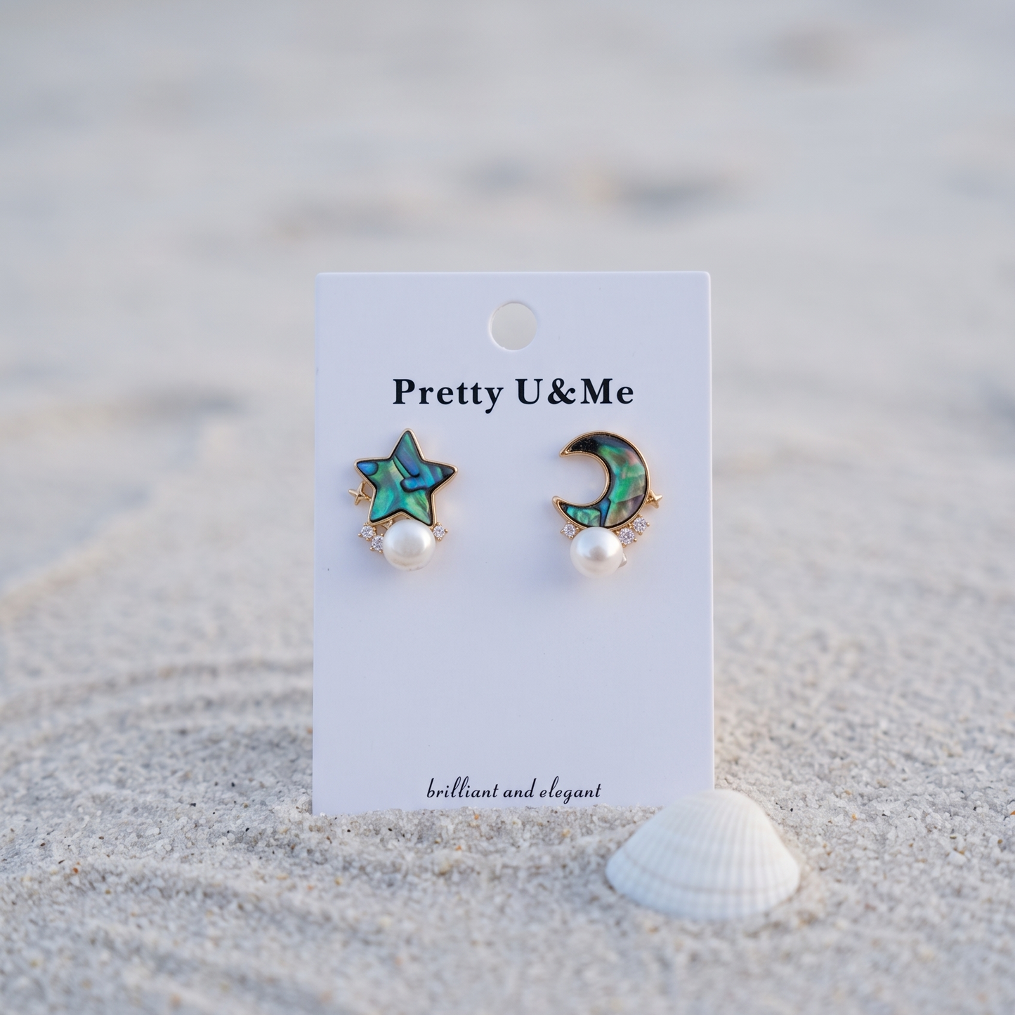 A pair of Pretty U&Me earrings displayed on a white card stand on a sandy beach. The earrings feature a turquoise and gold star on one post and a crescent moon on the other, each with a small white pearl dangling below.