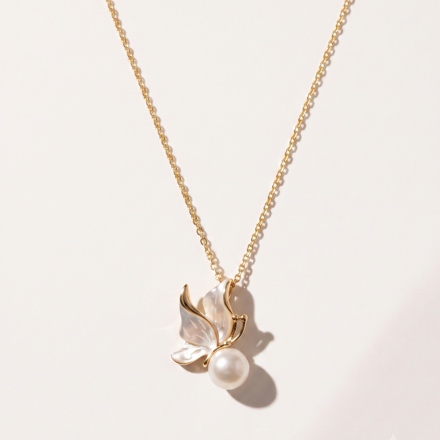 A delicate gold chain necklace featuring a pendant with mother-of-pearl butterfly wings and a single pearl at the center, displayed on a clean, light beige background.