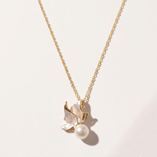 A delicate gold chain necklace featuring a pendant with mother-of-pearl butterfly wings and a single pearl at the center, displayed on a clean, light beige background.