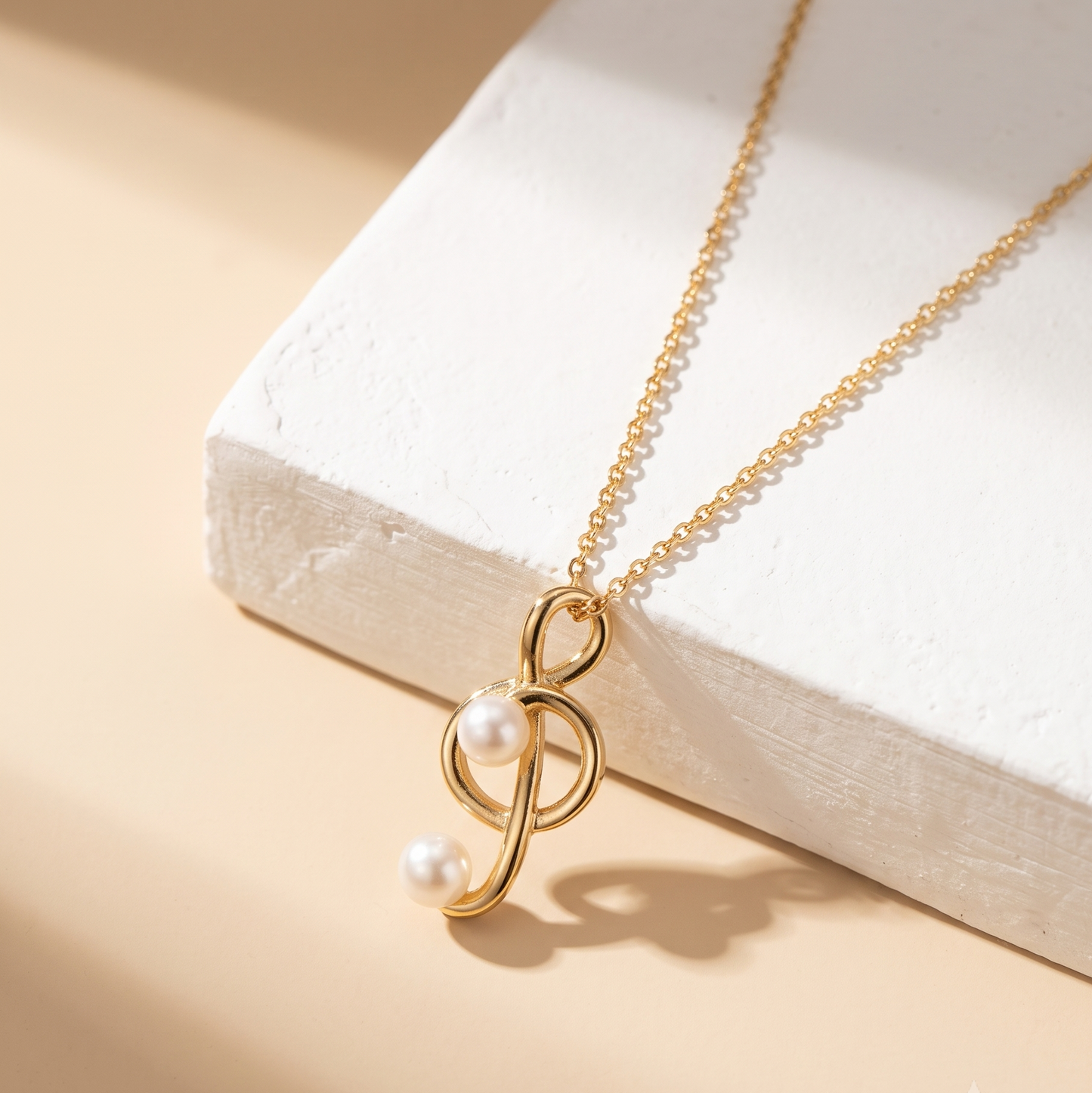 A gold chain necklace with a stylized infinity symbol pendant featuring two small white pearls, displayed on a textured white block against a warm beige background.