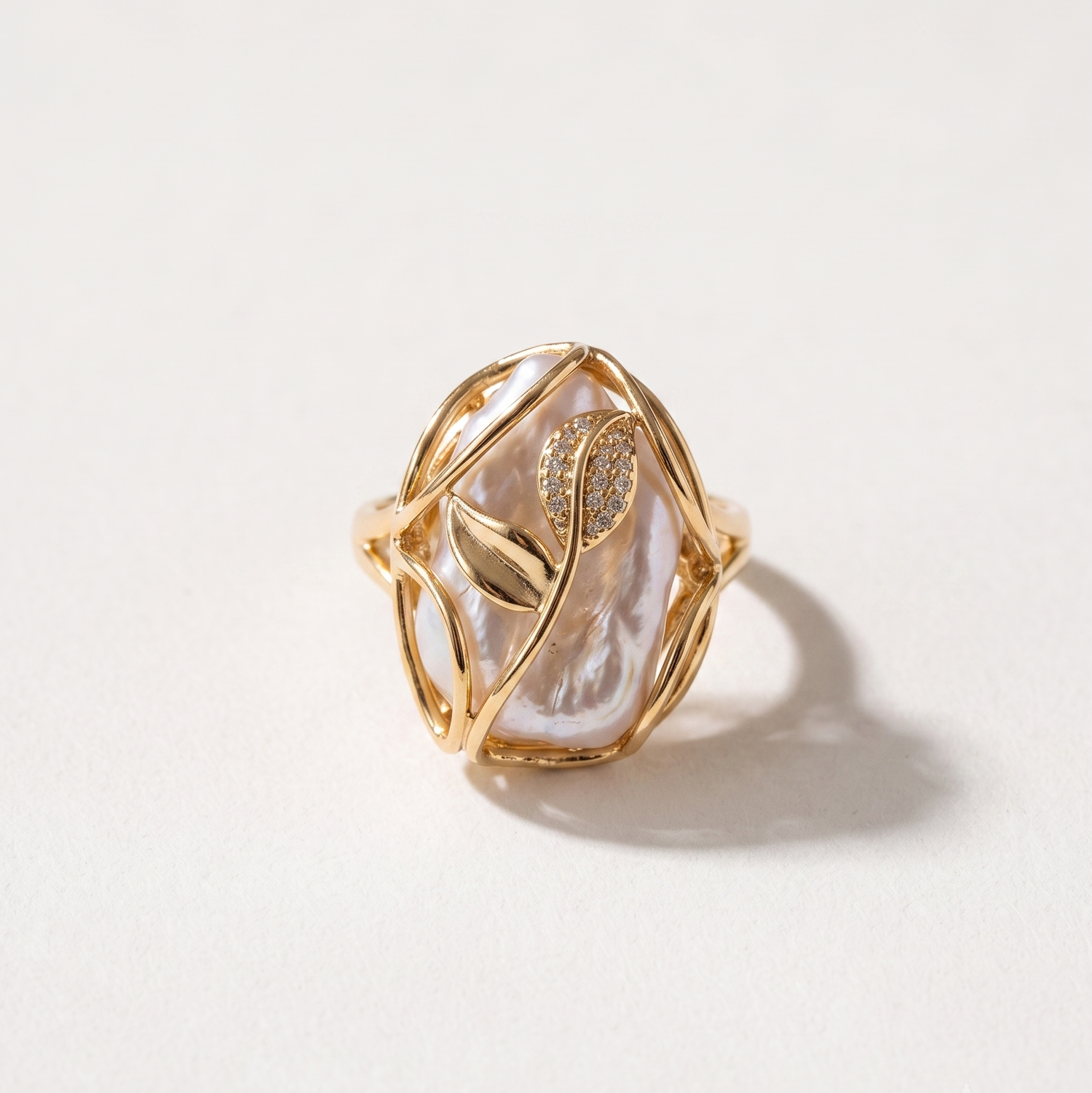 A gold ring featuring a large, organic-shaped white pearl with a gold leaf motif and small clear stones, set against a plain off-white background.