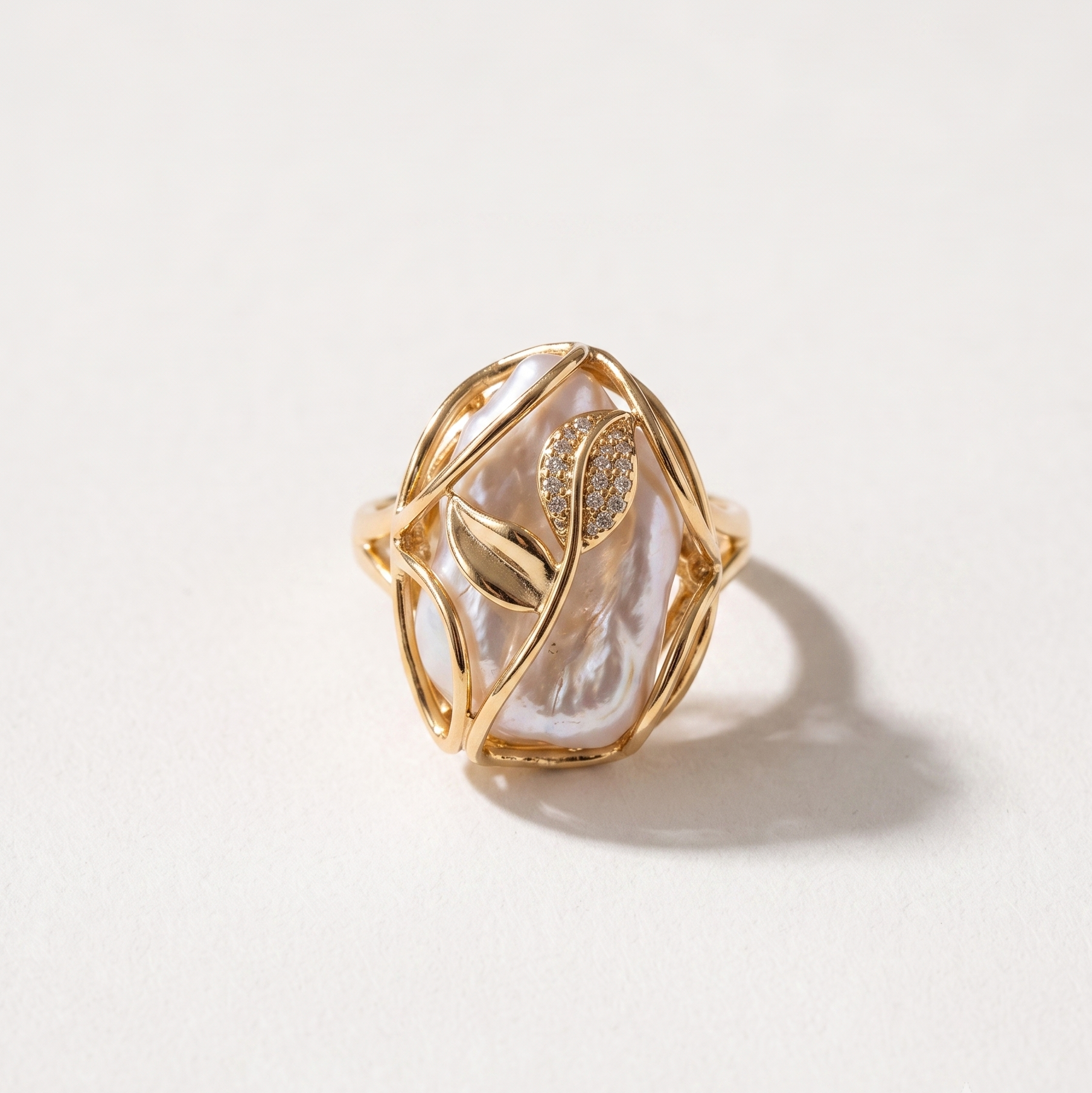 A gold ring featuring a large, organic-shaped white pearl with a gold leaf motif and small clear stones, set against a plain off-white background.
