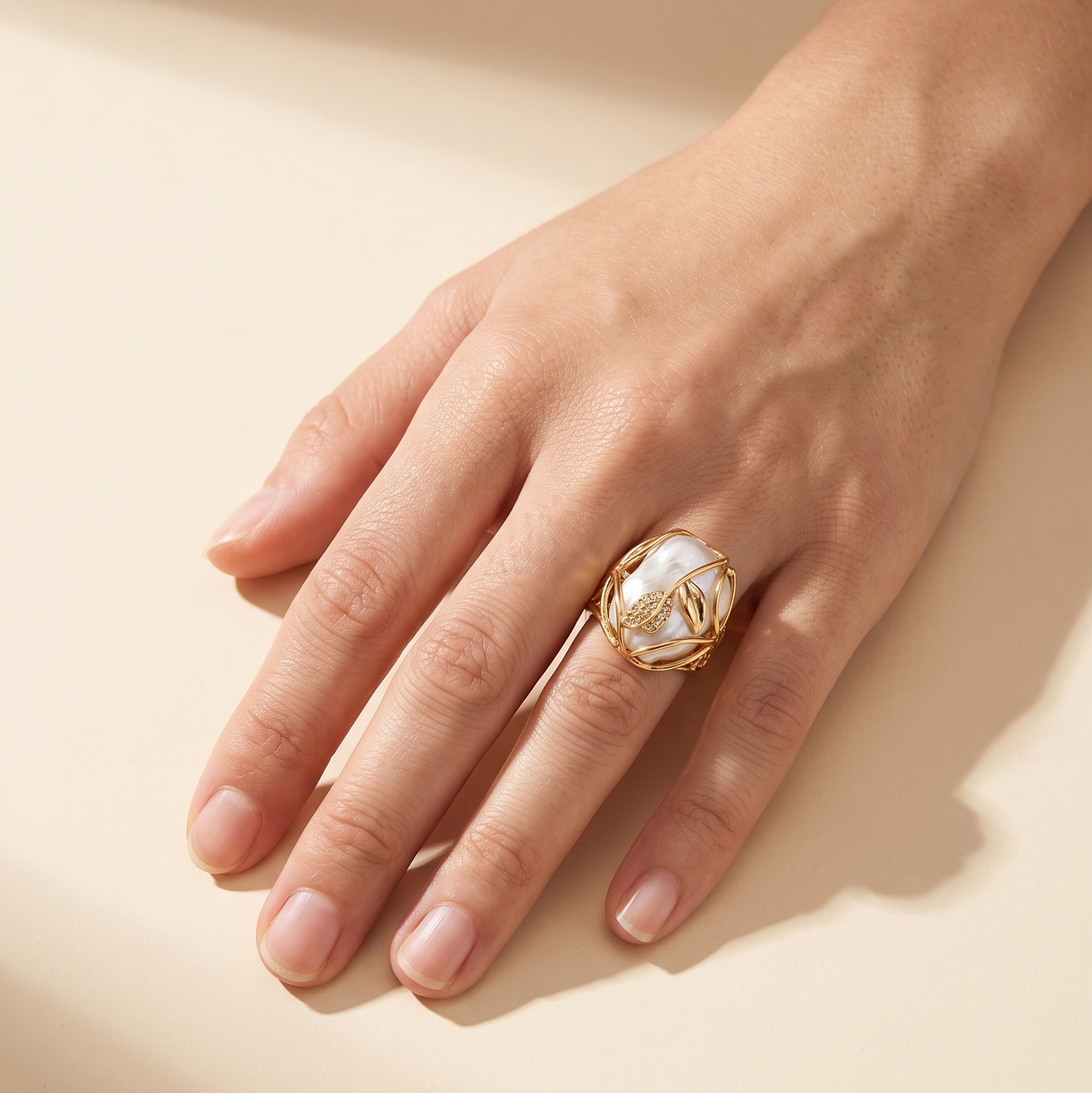 A gold ring with a large white floral inlay, worn on a hand against a warm beige background.