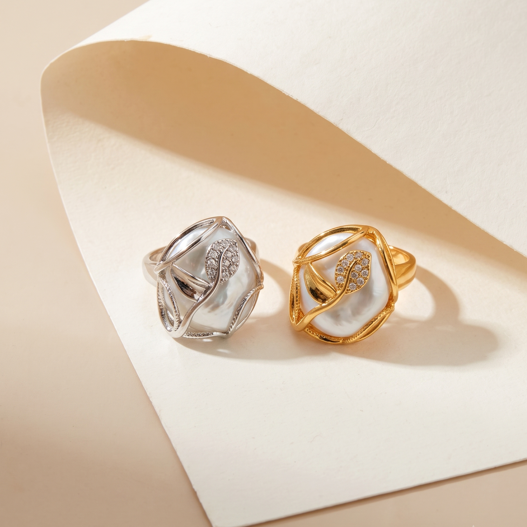 Two sculptural rings displayed on a folded cream paper surface against a warm beige background. One ring is silver-toned with a small clear stone cluster, and the other is gold-toned with a matching stone cluster.