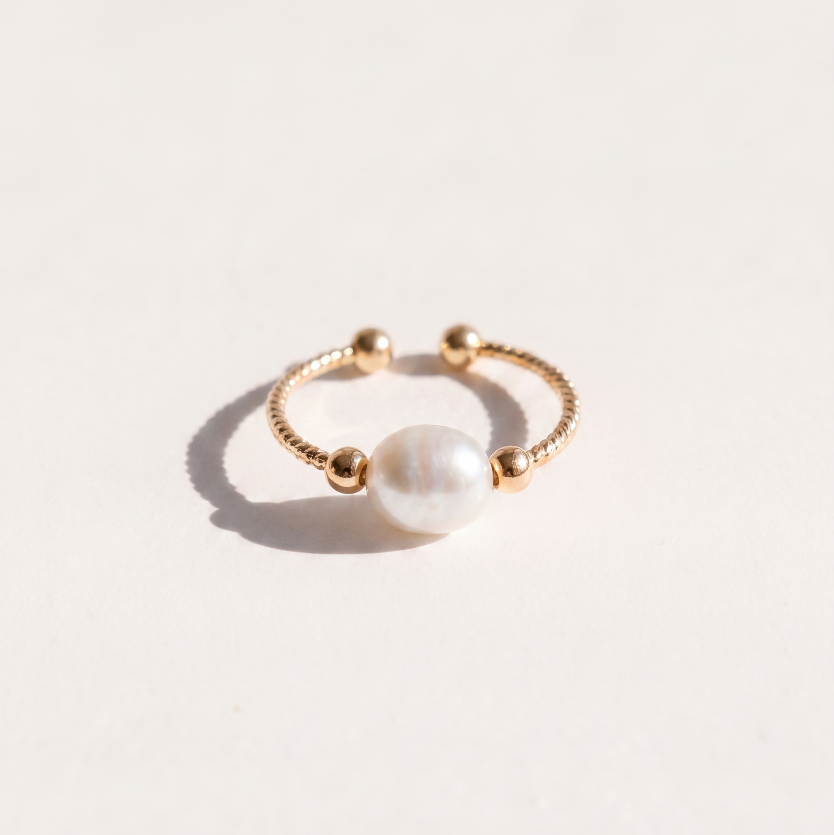 A delicate gold ring featuring a central white pearl bead, photographed on a clean, light off-white background with a soft shadow.