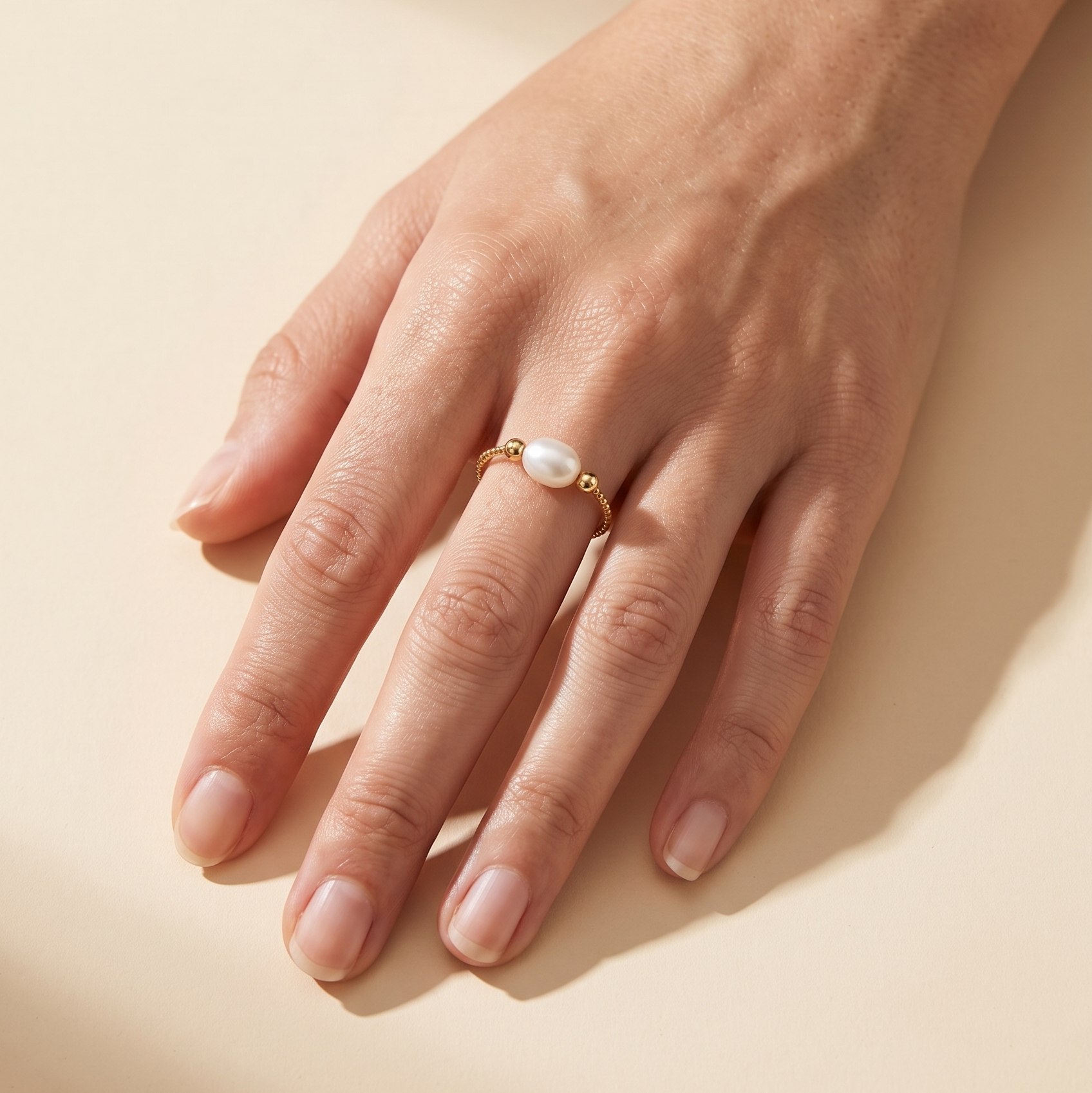 A delicate gold ring with a small white pearl center, worn on a person's ring finger against a warm beige background.