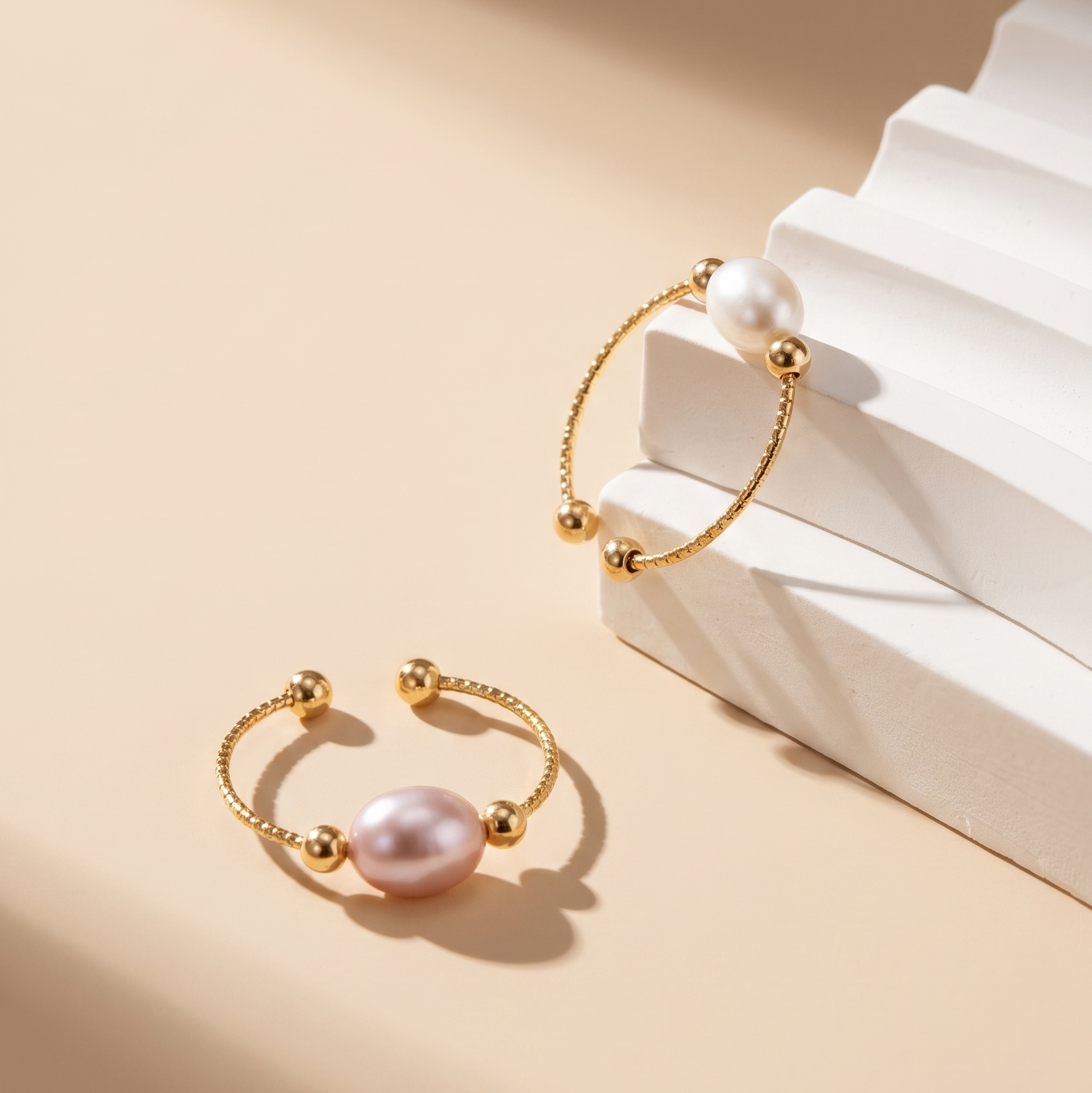 Two gold-toned open cuff bracelets with large pearl-like beads, styled on a beige background with white geometric blocks.