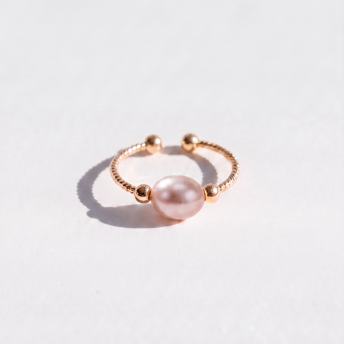 A delicate rose gold ring featuring a central light pink pearl bead and small gold-toned beads on the band, photographed on a clean white surface with a soft shadow.