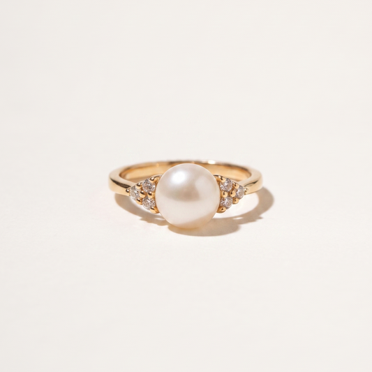 A gold ring featuring a large white pearl center stone and small clear accent stones on either side of the band, photographed on a clean off-white background.