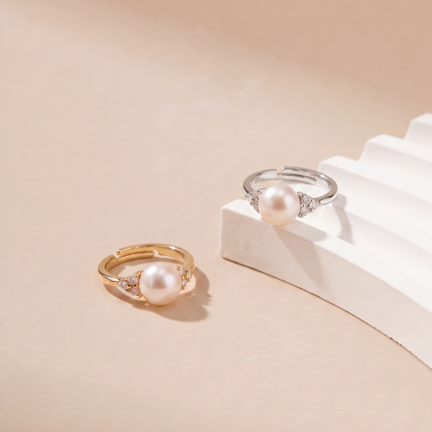 Two pearl rings, one in yellow gold and one in silver, displayed on a beige background with a white stepped prop.