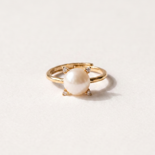 A gold ring featuring a round white pearl held in a four-prong setting with tiny accent stones, photographed on a clean white background.