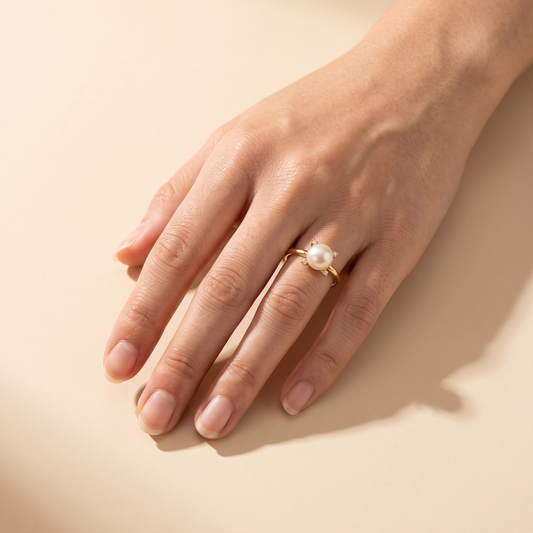 A close-up of a person's hand resting on a warm beige surface, wearing a delicate gold ring with a single round white pearl.