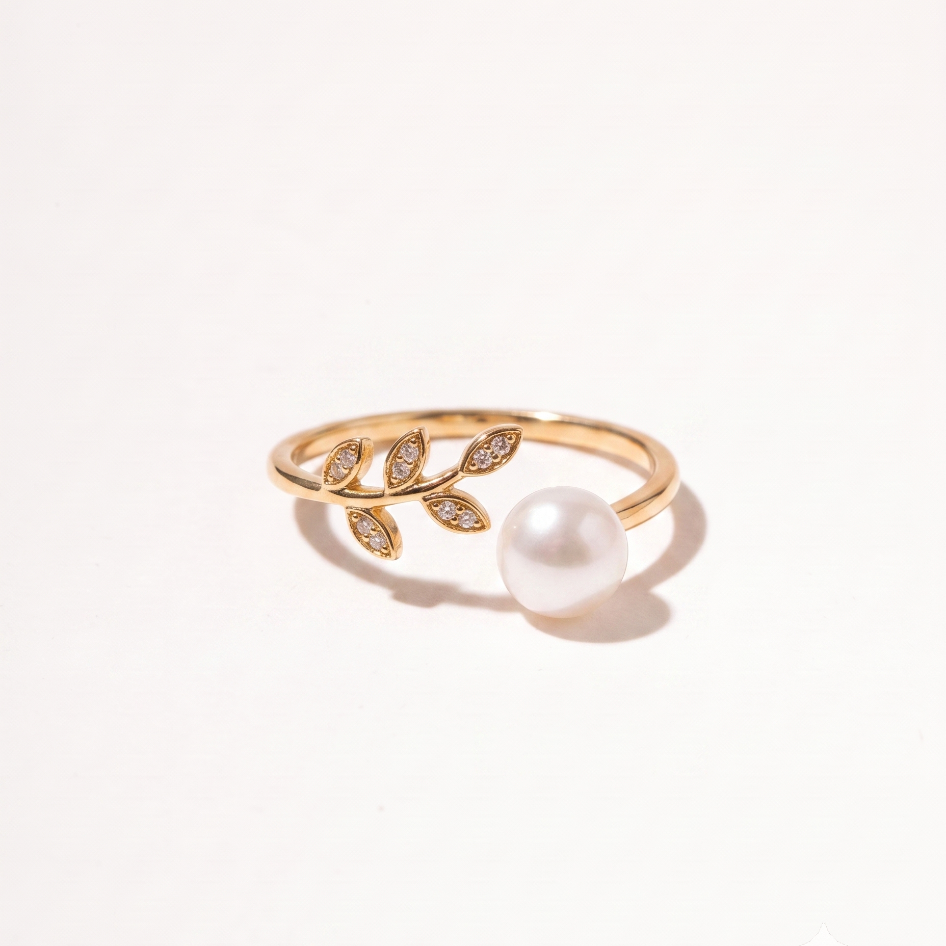 A gold ring featuring a leafy branch design with small pavé-set clear stones and a single white pearl, photographed on a clean white background.