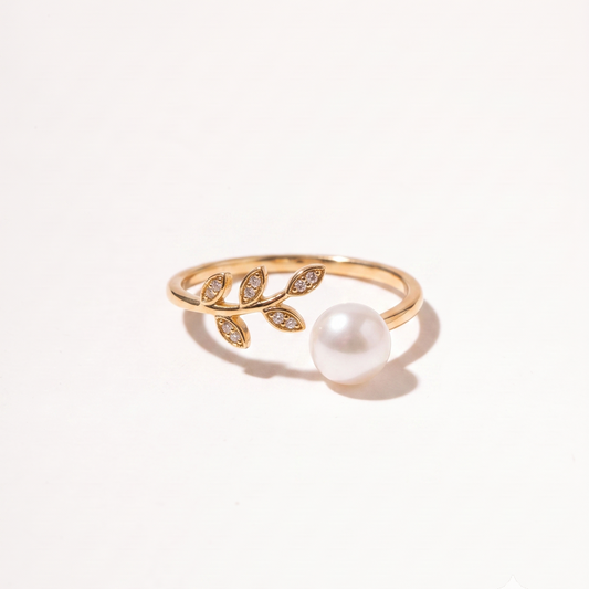 A gold ring featuring a leafy branch design with small pavé-set clear stones and a single white pearl, photographed on a clean white background.