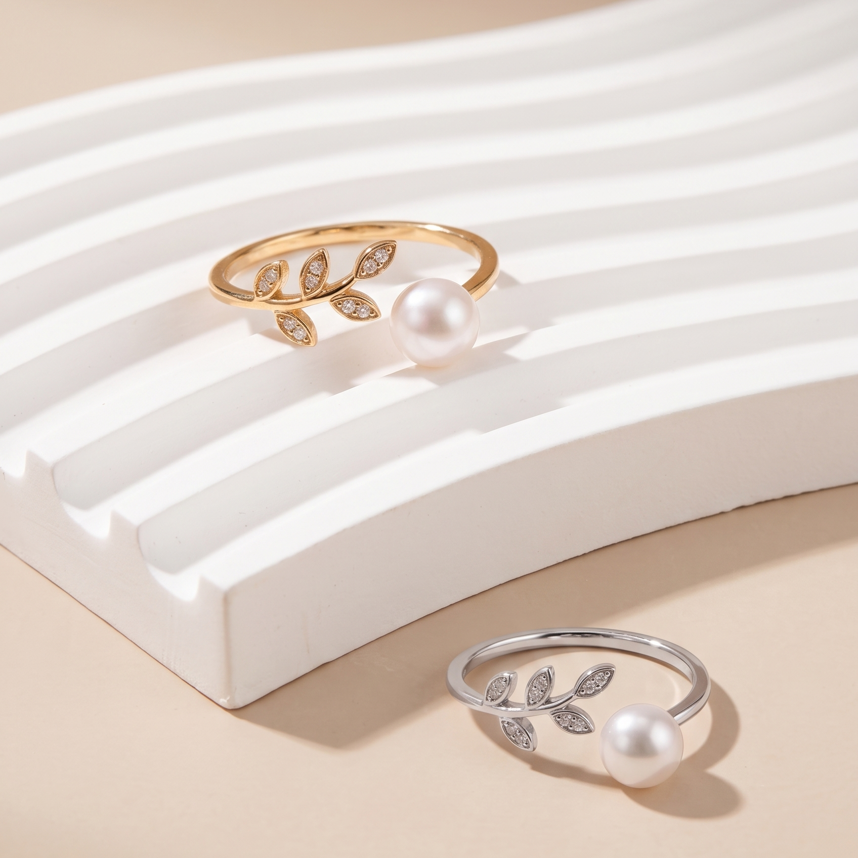 Two leaf-shaped open ring earrings with white pearls and small clear stones, shown on a beige background with a white curved display riser. One ring is gold-toned and one is silver-toned.