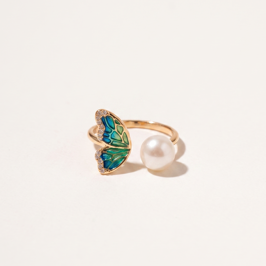 A gold butterfly ring with green and blue enamel detailing and a white pearl, photographed on a clean white background.