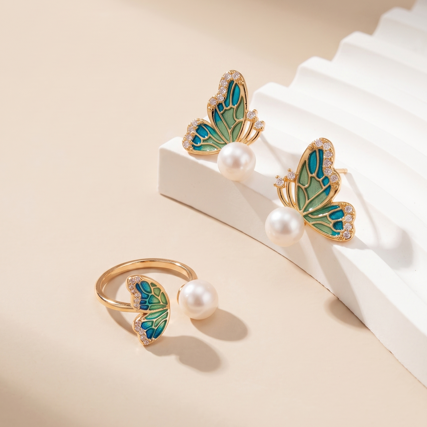 Gold butterfly stud earrings and a matching butterfly ring, each with a white pearl, displayed on a beige background with white geometric blocks.
