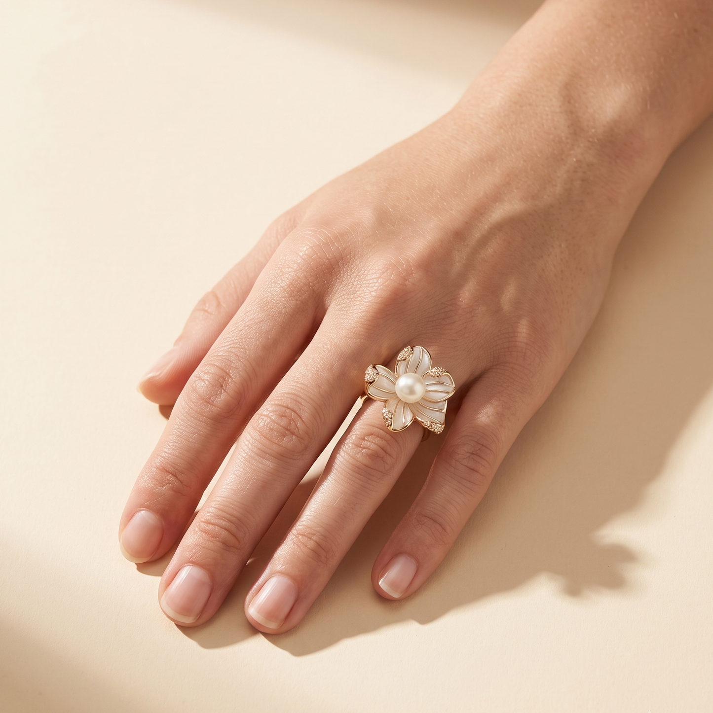 A hand wearing a delicate flower-shaped ring made of white pearls and small gold-toned metal accents, photographed on a warm beige background with soft shadows.