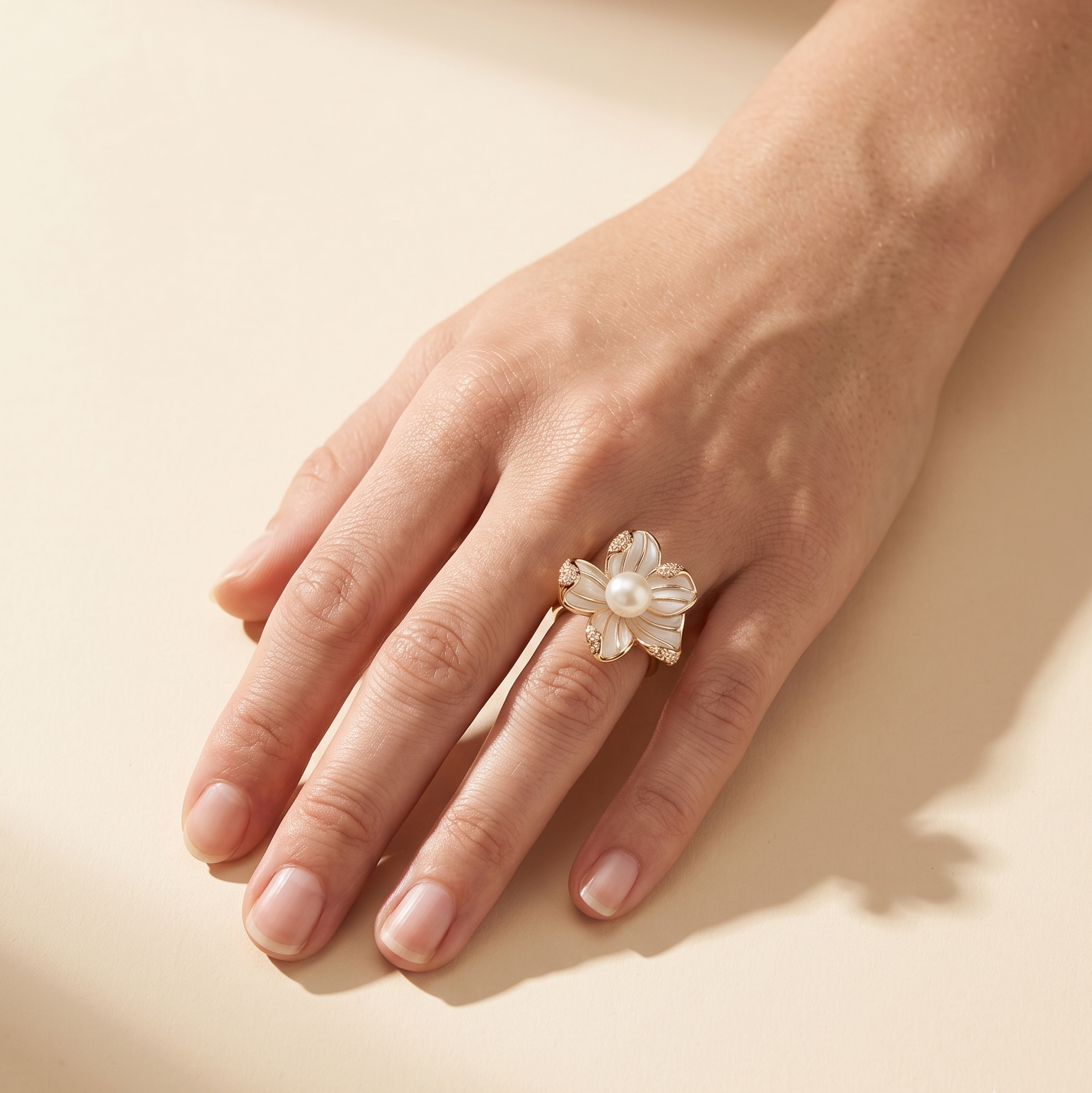 A hand wearing a delicate flower-shaped ring made of white pearls and small gold-toned metal accents, photographed on a warm beige background with soft shadows.