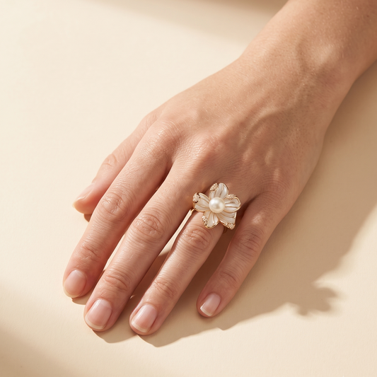 A hand wearing a delicate flower-shaped ring made of white pearls and small gold-toned metal accents, photographed on a warm beige background with soft shadows.