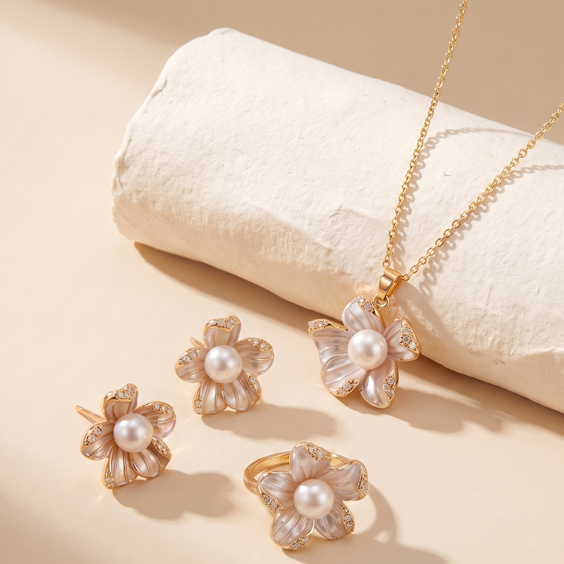 A gold jewelry set featuring a flower pendant necklace, a matching flower ring, and two matching flower stud earrings, each with a central white pearl and clear rhinestone petals, displayed on a beige background with a textured cream cylinder.