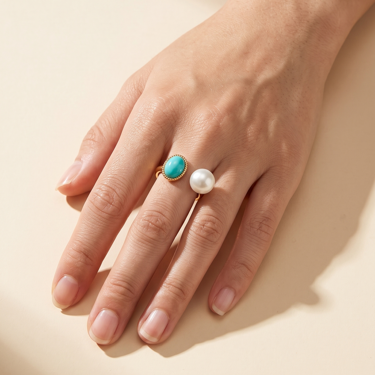 A close-up of a hand wearing two rings: a gold ring with a round turquoise stone and a gold ring with a white pearl, set against a warm beige background with soft shadows.