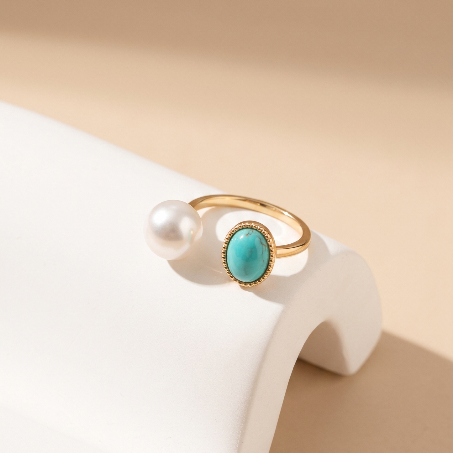 A gold open-band ring with a round turquoise stone and a dangling white pearl, displayed on a white curved jewelry stand against a warm beige background.